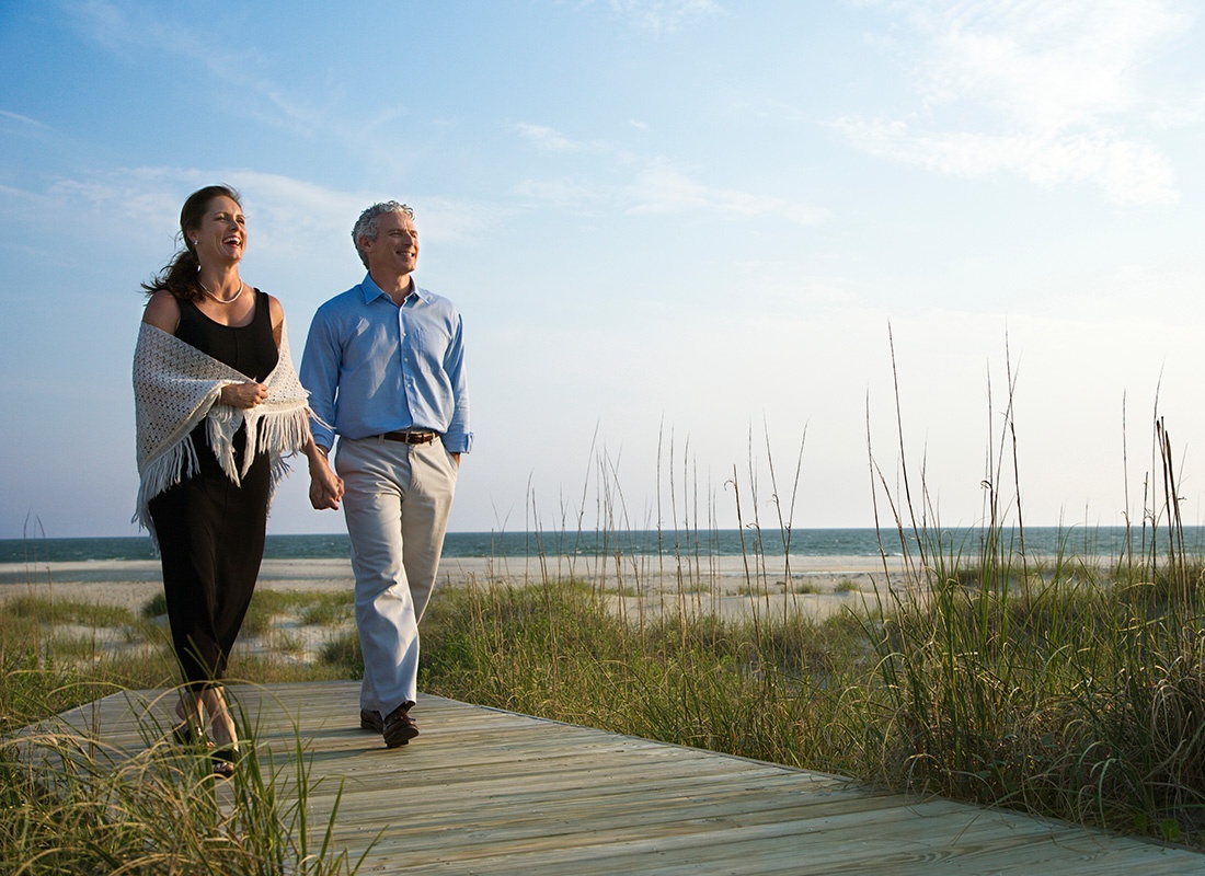 Employee Benefits - Older Couple Laugh and Walk Together on a Boardwalk by the Beach During Sunrise